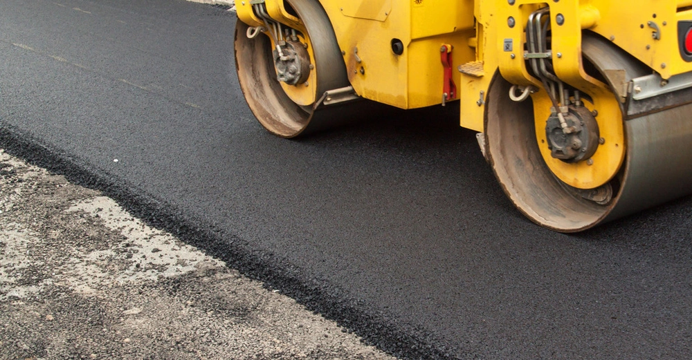 es wird asphaltiert: gelbe kleine Straßenwalze planiert frische, tiefschwarze Schwarzdecke, links daneben noch geschotterter Unterboden zu sehen