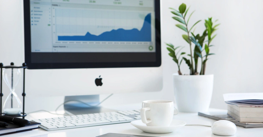 Ein Computermonitor zeigt ein Diagramm, eine Sanduhr und eine Tasse befinden sich auf einem Schreibtisch in einem hellen Büro mit Pflanzen.