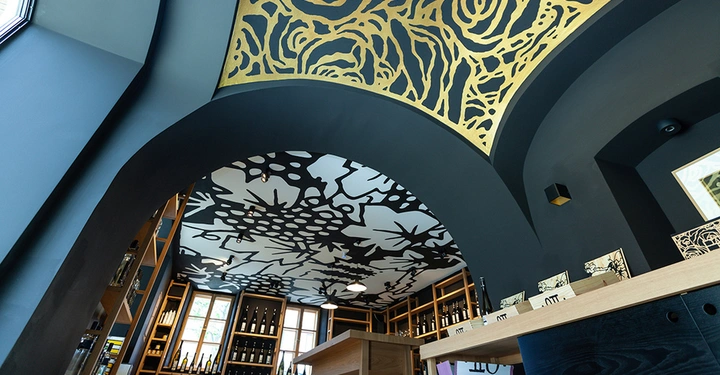 Galerie-Bild 13: Goldene Deckenornamente erstrecken sich über einem Raum mit Flaschenregalen und Fenstern während Licht von außen durch die Fenster eindringt durch eine braune hölzerne Theke im Vordergrund.