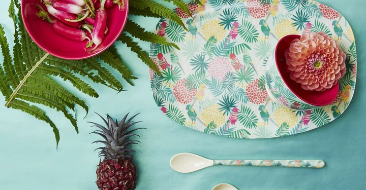 Galerie-Bild 5: Roter Teller mit Radieschen und blühender rosa Blume in einer roten Schale auf einem gemusterten Tablett auf einem türkisfarbenen Hintergrund umgeben von Farnblättern und einer Ananas daneben zwei weiße Löffel