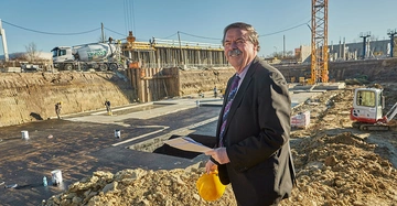 Mann hält Dokumente und Bauhelm lächelt baustellenartige Umgebung mit Betonierarbeiten und Baukran zahlreiche Arbeiter und Fahrzeuge wie Zementmischer und Bagger sind im Hintergrund aktiv