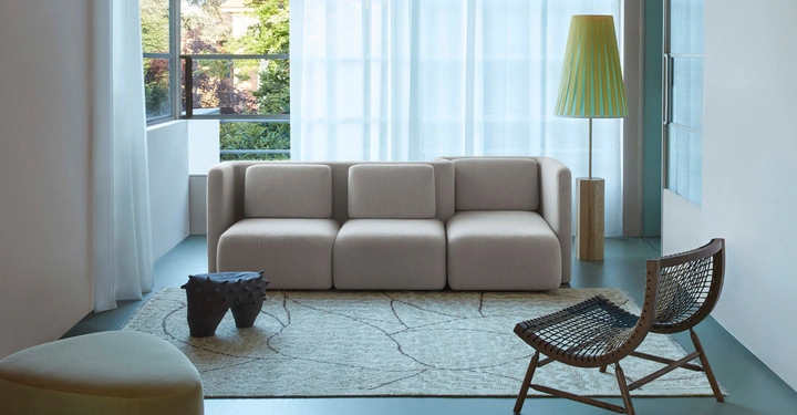 Beiges Sofa steht auf einem gemusterten Teppich vor großen Fenstern. Eine Stehlampe und zwei Stühle umgeben das Sofa in einem hellen Raum.