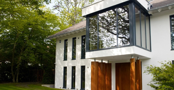 Großes modernes Haus mit weißer Fassade und großen Glasfenstern steht in einem grünen Garten umgeben von Bäumen und Rasenfläche