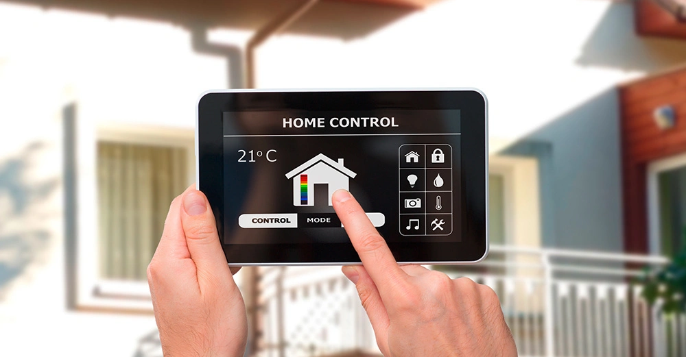 Eine Hand hält ein Tablet, das eine Home Control-App zeigt, während ein Finger auf das Display tippt 21°C, HOME CONTROL. Im Hintergrund ist ein modernes Haus zu sehen.