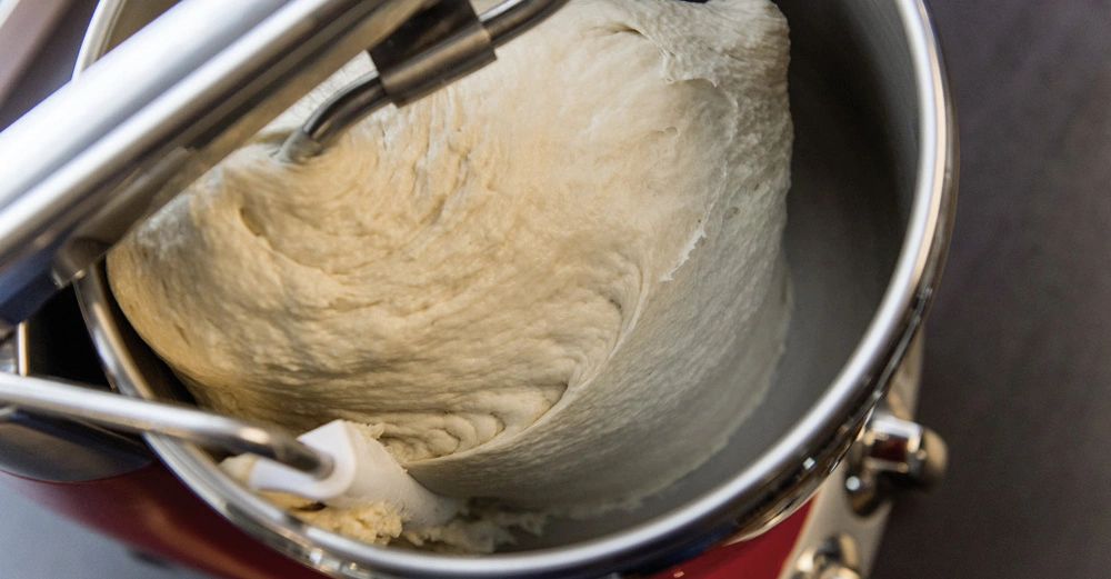 Ein Teig wird in einer Küchenmaschine geknetet. Die Metall-Schüssel und der Knethaken sind sichtbar. Die Küchenmaschine steht auf einer dunklen Arbeitsfläche in einem hellen Raum.