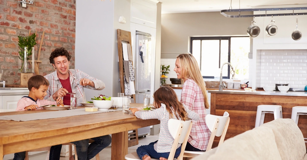Eine Familie sitzt an einem Holztisch und isst zusammen in einer modernen Küche mit weißen Wänden und Holzregalen. Der Vater spricht während die Mutter und Kinder essen.