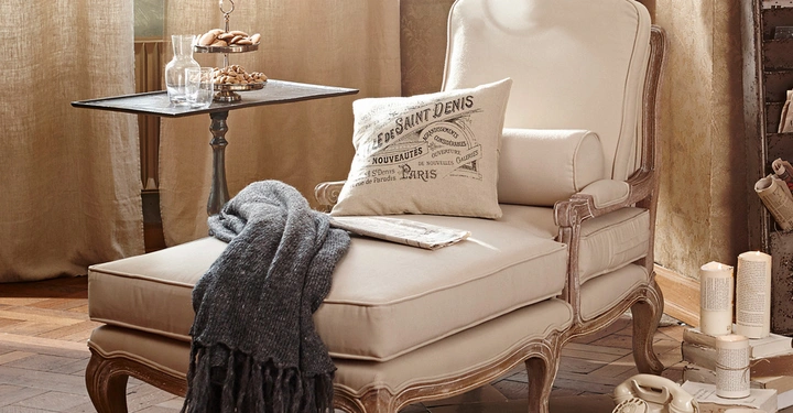 Galerie-Bild 5: Beiger Sessel und Ottoman mit grauer Decke und Kissen sitzend in einem gemütlichen Raum vor beige Vorhängen mit Beistelltisch voller Gebäck und alterm Telefon auf dem Holzboden. Text auf dem Kissen: LE DE SAINT DENIS.