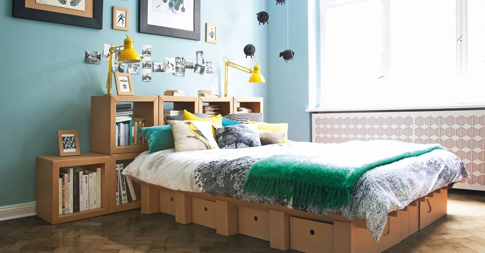 Ein Bett mit bunten Kissen steht in einem Schlafzimmer mit einem Parkettboden. Im Hintergrund sind Regale mit Büchern, gelben Lampen und Fotos an der blauen Wand.