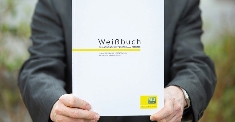 Ein Mann in einem Anzug hält ein weißes Buch mit dem Titel "Weißbuch Der Gemeinschaftsmarke ALU-Fenster Eine Entscheidungshilfe für Planer Architekten und Bauherren" im Freien.
