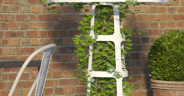 Weißer Pflanzständer umschlingt mit grünen Blättern von einer Kletterpflanze steht auf einem Betonboden vor einer Ziegelwand neben einem Metallstuhl und einem großen Blumentopf mit einem Busch