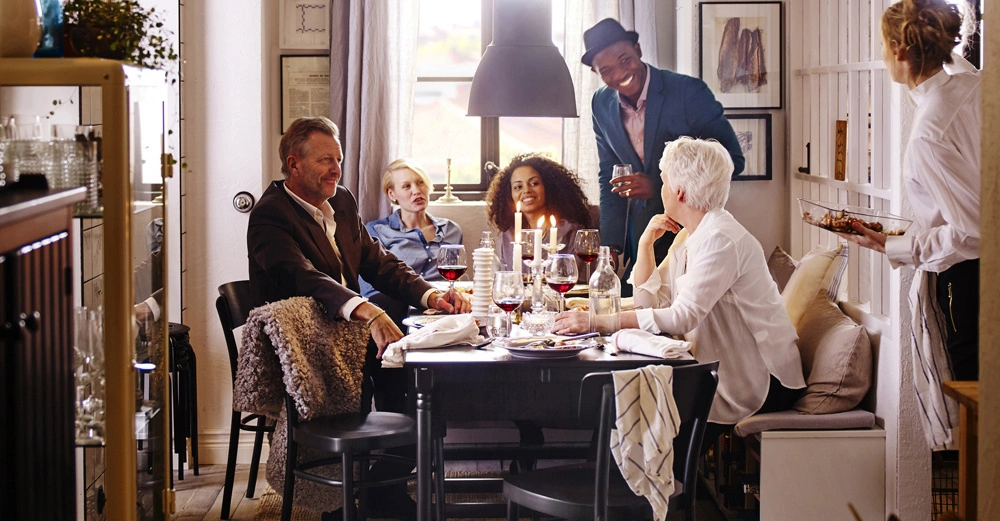 Menschen sitzen um einen gedeckten Esstisch und unterhalten sich in einem gemütlichen Esszimmer mit Kerzenleuchtern und dekorativen Bildern an den Wänden. Eine Kellnerin bringt Essen.