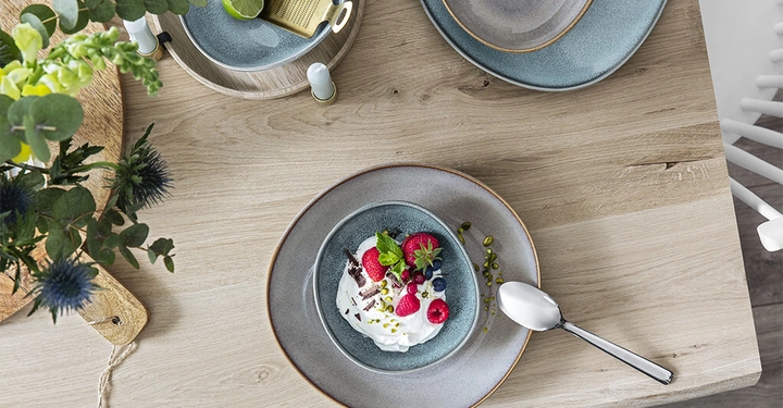 Galerie-Bild 2: Vogelperspektive von Platzteller mit Steingutschüssel drauf in Blaugrau von Villeroy & Boch. In der Schüssel Joghurt mit Erdbeeren, Blüten und goldenem Löffel. 