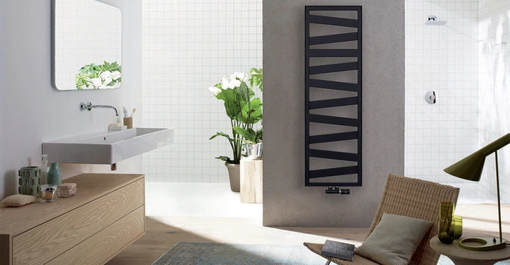 Schwarzer Handtuchheizkörper hängt an grauer Wand moderner hell eingerichteter Raum mit Waschbecken Spiegel Pflanzen Holzkommode geflochtenem Stuhl grünem Kissen und Lampe daneben.