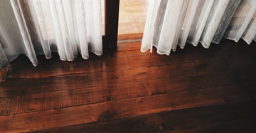 Weiße Gardinen hängen vor einer Glastür, die zu einem Raum mit einem polierten Holzboden führt, der Sonnenlicht reflektiert.