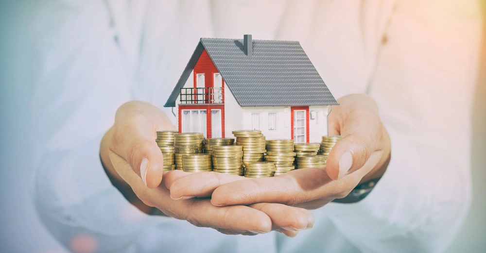 Eine Person hält ein Miniatur-Einfamilienhaus und gestapelte Münzen in ihren Händen. Im Hintergrund ist ein unscharfer heller Bereich zu sehen.