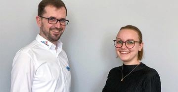 Ein Mann mit weißem Hemd und eine Frau in schwarzem Pullover lächeln in die Kamera in einem neutralen Innenraum. Das Hemd hat Logos: Saint-Gobain und ein unlesbares bläuliches Logo