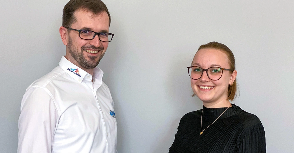 Ein Mann mit weißem Hemd und eine Frau in schwarzem Pullover lächeln in die Kamera in einem neutralen Innenraum. Das Hemd hat Logos: Saint-Gobain und ein unlesbares bläuliches Logo
