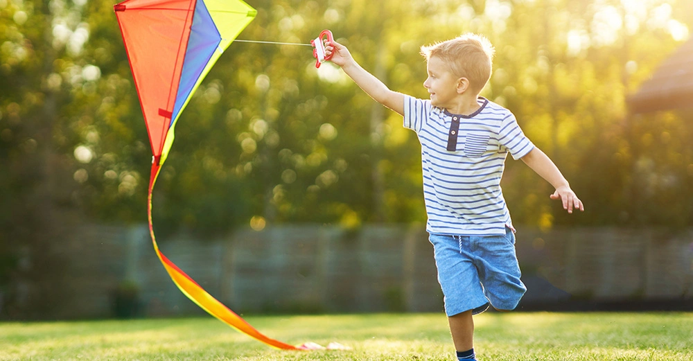 kleiner Junge in blauer Shorts und gestreiftem Shirt läuft glücklich mit buntem Dreiecks-Drachen durch den Garten, Sonne scheint