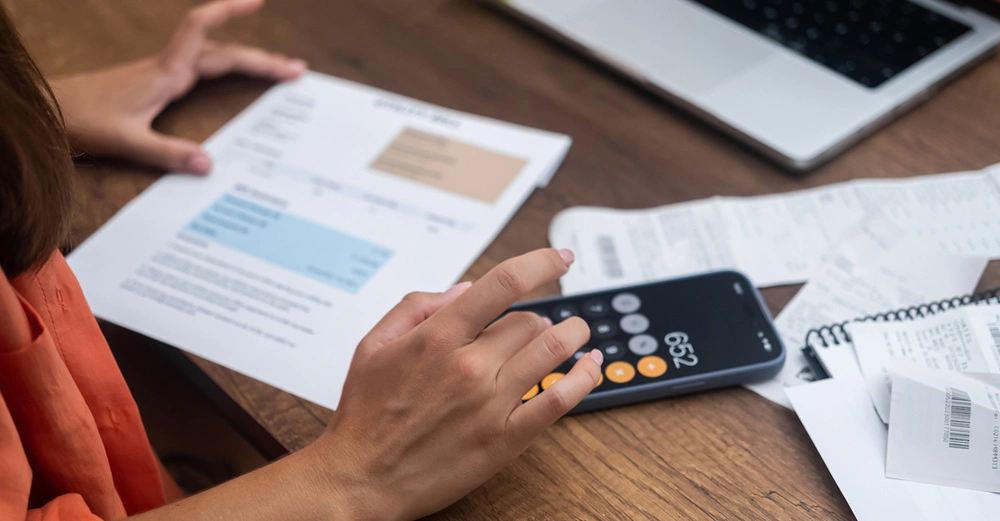 Frau berechnet etwas auf Taschenrechner, auf dem Tisch liegen Unterlagen, ein Laptop und Kugelschreiber.