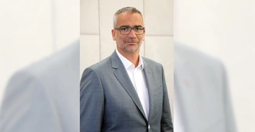 Mann trägt grauen Anzug und weißes Hemd steht vor grauer Wand im Innenbereich