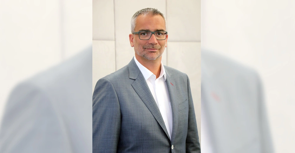 Mann trägt grauen Anzug und weißes Hemd steht vor grauer Wand im Innenbereich