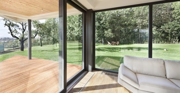 Eine helle weiße Couch steht in einem Raum mit großen bodentiefen Fenstern die zum grünen Garten und einer Holzterrasse zeigen im Hintergrund sind Bäume und Gartenmöbel zu sehen.