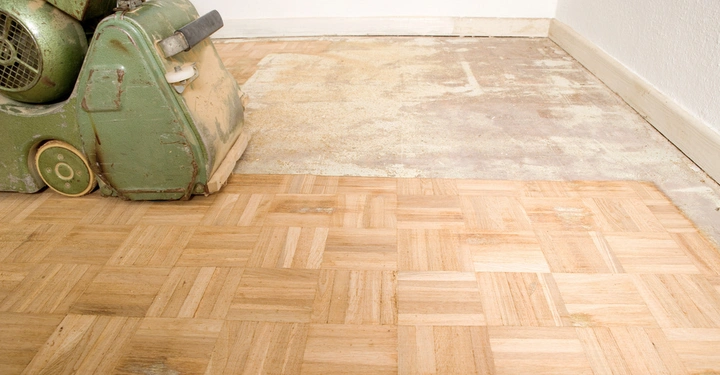 Eine grüne Schleifmaschine schleift einen Holzparkettboden in einem Raum mit weißer Wand und Fußleiste. Links glattes Holz; rechts ungeschliffen.