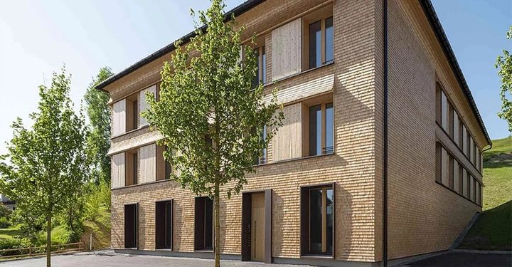 Ein dreistöckiges Gebäude aus Holz steht still zwischen zwei Bäumen im Vordergrund in einer grünen Umgebung mit Hügeln im Hintergrund unter einem blauen Himmel