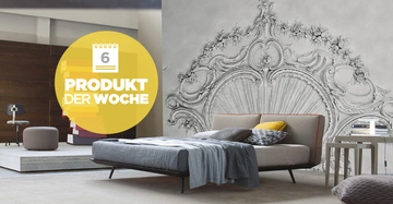 Ein Bett mit grau bezogener Bettwäsche und dekorativen Kissen steht vor einer kunstvollen Wandmalerei in einem modernen Schlafzimmer. Linke Seite steht ein Stuhl und ein Hocker. Text: 6 PRODUKT DER WOCHE