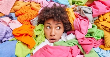 Person schaut überrascht zwischen bunten Kleidungsstücken hervor Kontext ist eine Fülle farbenfroher Stoffe die die Person umgeben und teilweise verdecken