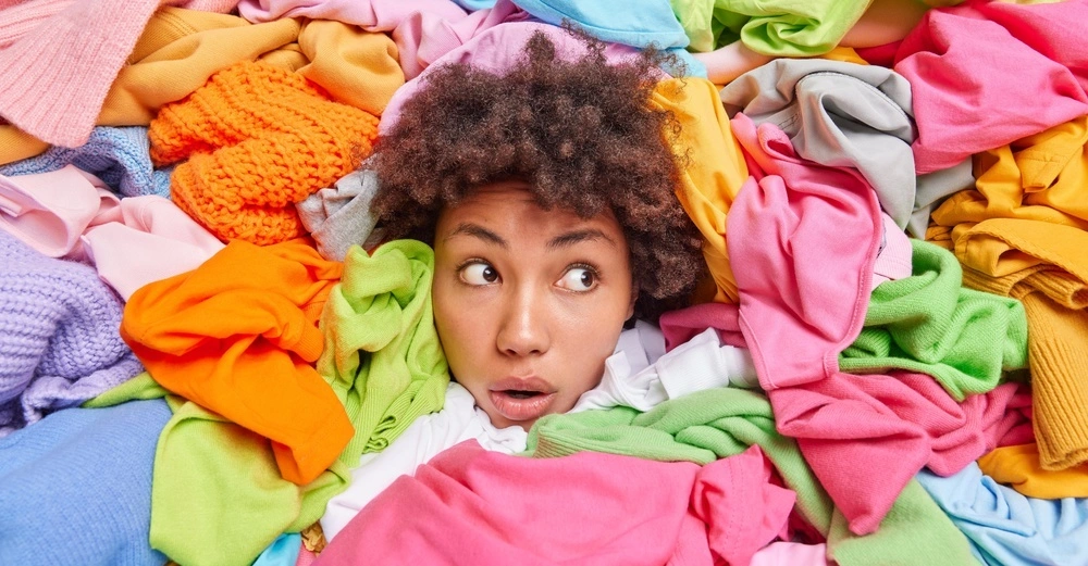 Person schaut überrascht zwischen bunten Kleidungsstücken hervor Kontext ist eine Fülle farbenfroher Stoffe die die Person umgeben und teilweise verdecken