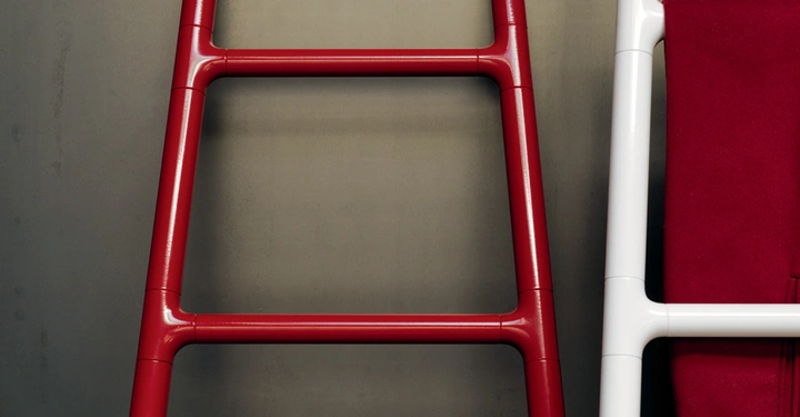 Rote glänzende Leiter lehnt an einer grauen Wand während eine weiße Leiter mit einem roten Tuch daneben steht.