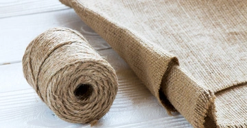 Eine Rolle Jutegarn liegt auf einer weißen Holzoberfläche neben einem teilweise ausgerollten Stück Sackleinen.