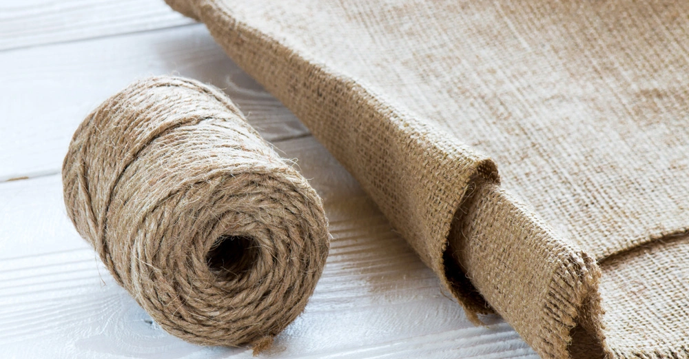 Eine Rolle Jutegarn liegt auf einer weißen Holzoberfläche neben einem teilweise ausgerollten Stück Sackleinen.