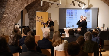 Eine Frau und zwei Männer von DenkMalNeo stehen auf einer Bühne vor Publikum.