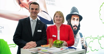 Zwei Personen stehen lächelnd an einem Messestand mit Broschüren und Snacks Eine bunte Wand mit Abbildungen von Menschen im Hintergrund Text: Einfach Flexibel und teilweise verdeckt