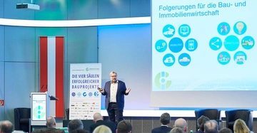 Ein Mann hält einen Vortrag vor einer Gruppe von Menschen. Er steht neben einem Rednerpult und zeigt auf eine Projektionsleinwand mit den Texten "Die vier Säulen erfolgreicher Bauprojekte" und "Folgerungen für die Bau- und Immobilienwirtschaft". Der Raum ist modern eingerichtet.