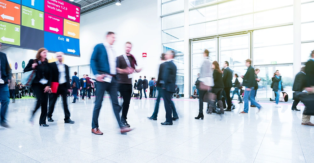 Menschen gehen in einer hellen Messehalle herum. Ein großes Schild zeigt Wegweiser mit Texten wie Debate Hall Seminars 1.2.3 Work Labs VIP Lounge Press Center Market Place 1 and 2 Exit.