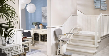 Treppenlift an einer weißen Treppe fährt nach oben in einem modernen, hellen Raum mit Pflanzen; im Hintergrund sind Körbe und Weinregale auf hellen Möbeln zu sehen.