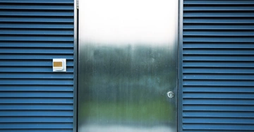 Metalltür steht still umgeben von blauen Lamellenwänden in einer modernen Architekturumgebung mit einem kleinen weißen Sicherheitskasten links von der Tür.