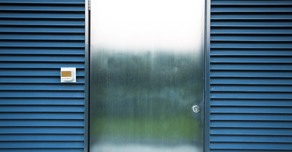 Metalltür steht still umgeben von blauen Lamellenwänden in einer modernen Architekturumgebung mit einem kleinen weißen Sicherheitskasten links von der Tür.