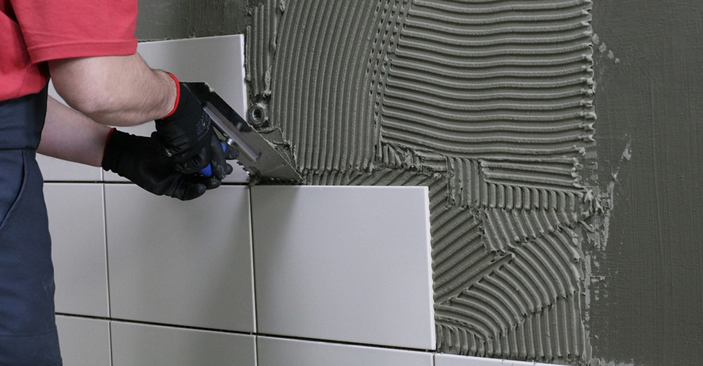 Eine Person in einem roten Shirt und schwarzen Handschuhen verlegt beige Fliesen an einer Wand mit grau strukturiertem Fliesenkleber in einem Innenraum.