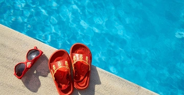 Rote Sandalen und rote Sonnenbrille liegen auf einem Steg neben einem blauen Schwimmbecken.