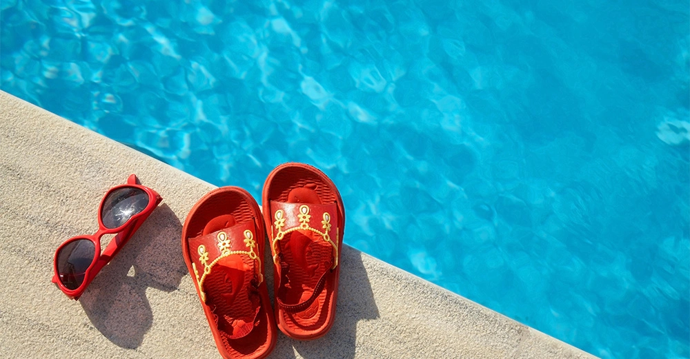 Rote Sandalen und rote Sonnenbrille liegen auf einem Steg neben einem blauen Schwimmbecken.