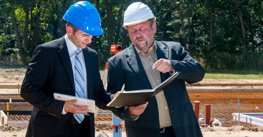 zwei Anzugträger mit Bauhelmen haben Unterlagen und Ordner in der Hand und stehen auf der Baustelle.