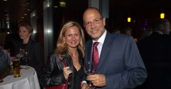 Galerie-Bild 82: Ein Mann und eine Frau halten Getränke und lächeln in die Kamera Partyumgebung mit mehreren Personen im Hintergrund und Tischen mit Gläsern und Flaschen.