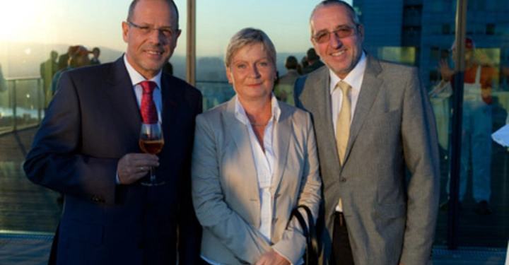 Galerie-Bild 5: Drei Personen posieren lächelnd in formeller Kleidung auf einer Dachterrasse mit Glasgeländern bei Sonnenuntergang vor einem modernen Glasgebäude. Eine Person hält ein Weinglas. Weitere Menschen sind im Hintergrund zu sehen.