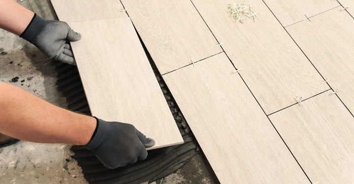 Hand mit Handschuhen platziert eine Fliese auf Boden Mörtel wird aufgetragen und Fliesen sind in gleichmäßigen Reihen mit Abstandhaltern in einem Raum positioniert.