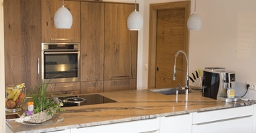Küchenbereich mit Holzschränken, Hängelampen und eingebautem Backofen im Holzschrank Handlungen sind nicht sichtbar; auf der Theke befinden sich Obst, Pflanzen, Herdplatte, Spüle und Kaffeeautomat.