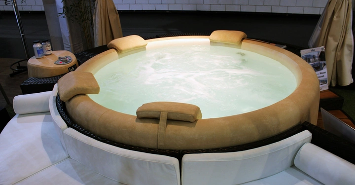 Galerie-Bild 11: Runder Whirlpool mit sprudelndem Wasser umgeben von beigen und weißen Polstersitzen im Hintergrund stehen beige Vorhänge und ein runder Tisch mit Flaschen und Bedienfeldern vor einer gefliesten Wand